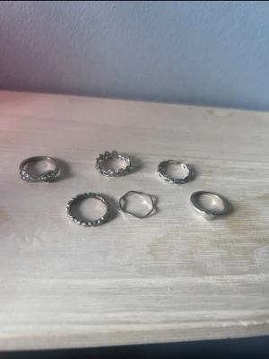Sats med sex silverfärgade ringar - Säljer ett set med sex olika ringar i silverfärg. Ringarna har varierande design, bland annat med pärldetaljer, tvinnad form och hjärtformad detalj. Passar perfekt för att mixa och matcha flera på samma hand. Materialet ser ut att vara metall.