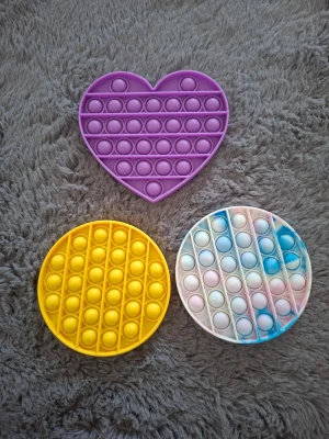 Tre pop it-leksaker i olika färger - Säljer tre pop it-leksaker i silikon: en lila hjärtformad, en gul rund och en rund i vitt, rosa och blått. Perfekta för att stressa ner eller bara ha kul med. Alla har bubblor att trycka på och är lätta att ta med sig.