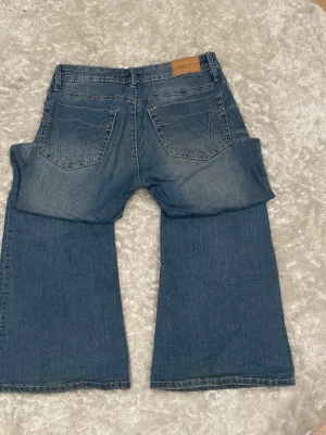 Blå bootcut jeans med utsvängda ben - Säljer ett par blå bootcut jeans med utsvängda ben och klassisk femficksmodell. Jeansen har en snygg tvätt med ljusa partier framtill och bakfickor med diskreta sömmar. Perfekta för dig som gillar retrostil och vill ha ett par bekväma jeans i bomull.