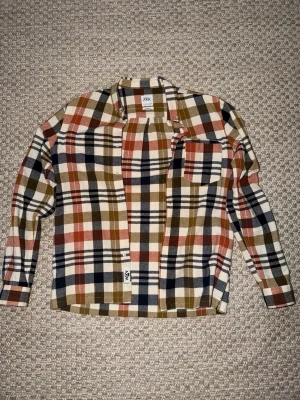 Rutig skjorta från Zara i höstfärger - Säljer en rutig skjorta från Zara i beige, brun, marinblå och rostorange. Skjortan har klassisk krage, bröstficka och knappar framtill. Tillverkad i mjuk bomull, perfekt för lager på lager och en chill look.