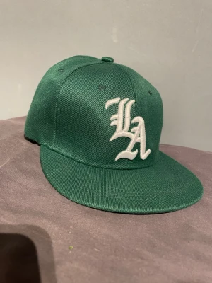 Grön snapback keps med LA-brodyr - Snygg grön snapback keps med vit LA-brodyr framtill. Klassisk platt skärm och justerbar baksida. Tillverkad i ett tåligt tygmaterial som ger en clean och sportig look.