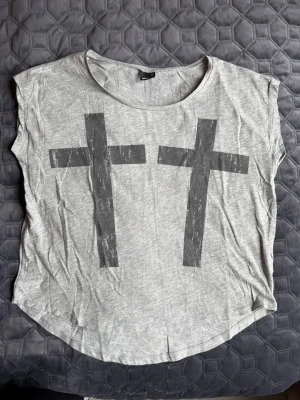 Grå t-shirt med tryck från Gina Tricot - Säljer en grå t-shirt från Gina Tricot med två stora svarta kors på framsidan. T-shirten har rund halsringning och är kortärmad med en loose passform. Perfekt för en avslappnad och edgy stil.