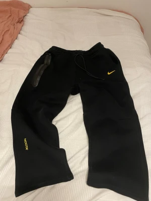 Svart Nike NOCTA hoodie med huva - Svart hoodie från Nike NOCTA med gul logga på bröstet och NOCTA-tryck på ärmen. Dragkedja framtill, två fickor med svarta detaljer och en skön huva. Perfekt för en streetwear-look och riktigt bekväm passform.