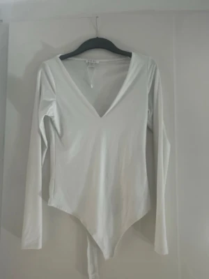 Vit långärmad body med V-ringning - Säljer en stilren vit body med lång ärm och djup V-ringning. Bodyn har en enkel och clean design, perfekt att matcha med olika outfits. Tillverkad i mjukt stretchigt material för skön passform.