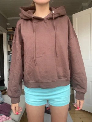 Brun hoodie med huva och dragsko - Mysig brun hoodie med huva och långa ärmar. Modellen är oversized med ribbade muddar vid ärmslut och nederkant. Hoodien har dragsko i halsen och är perfekt för en avslappnad stil. Tillverkad i mjukt material för extra komfort.