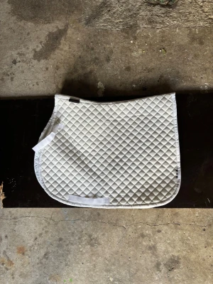 Vitt schabrak med rutmönster - Säljer ett vitt schabrak med klassiskt rutmönster. Schabraket har rundade hörn och kardborreband för enkel fastsättning på sadeln. Materialet är quiltat tyg som ger bra komfort till hästen. Perfekt till ridning och träning.
