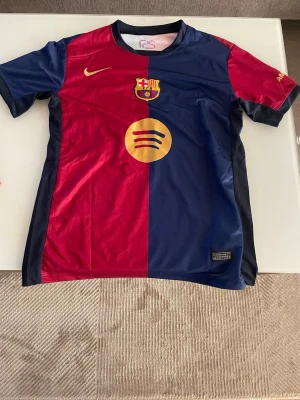 FC Barcelona matchtröja Lamine Yamal - Snygg FC Barcelona matchtröja i rött och marinblått med guldfärgade detaljer. Tröjan har klubbmärke, Nike-logga och Spotify-logga på framsidan samt Lamine Yamal och nummer 19 på ryggen. Tillverkad i lätt och ventilerande material, perfekt för fotbollsträning.