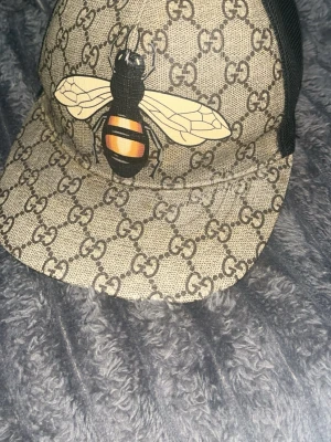 Gucci keps med bi och GG-mönster - Säljer en snygg Gucci keps med klassiskt beige GG-mönster och stor bi-detalj framtill. Kepsen har svart mesh baktill för extra ventilation och justerbar passform. Materialet är canvas framtill och mesh bak. Perfekt för dig som vill sticka ut med en unik accessoar.