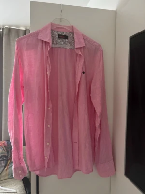 Rosa linneskjorta från Morris - Säljer en snygg rosa skjorta från Morris i linne. Skjortan har klassisk krage, knappar framtill och lång ärm. Insidan av kragen har ett paisleymönster i blått och grått som ger en extra detalj. Perfekt för dig som vill ha en fräsch och stilren look.