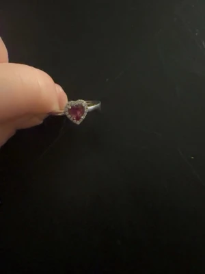 Silverring med hjärtformad röd sten - Säljer en söt silverfärgad ring med en hjärtformad röd sten i mitten, omgiven av små klara stenar som ger extra bling. Perfekt för dig som gillar romantiska och unika smycken. Ringen har en tunn, elegant ringband och passar till många olika stilar.