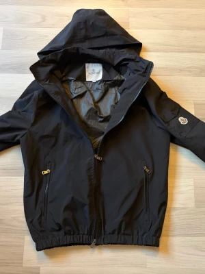 Svart vindjacka från Moncler - Snygg svart vindjacka från Moncler med huva som går att ta bort och dragkedja framtill. Jackan har två snedställda fickor med dragkedja och elastisk nederkant. Moncler-logga på ärmen ger en exklusiv touch. Tillverkad i lätt och tåligt material, perfekt för blåsiga dagar. Köpt av min vän vet ej om den är äkta men otroligt fint skick 