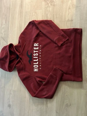 Vinröd hoodie från Hollister - Snygg vinröd hoodie från Hollister med broderad logga och texten 'Hollister California' på bröstet. Hoodien har huva med dragsko och en stor magficka. Mjuk och skön bomullsblandning, perfekt för chill dagar.