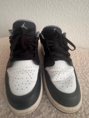 Svarta och vita Air Jordan 1 Low - Säljer ett par Nike Air Jordan sneakers i svart och vitt. Skorna har svart läder på tå och snörning, vit tåbox med perforeringar och vit sula. Ikoniska Jumpman-loggan syns på plösen. Perfekta för dig som gillar streetwear och sportig stil. Nopprigt i hälarna, men ingenting som syns utåt.