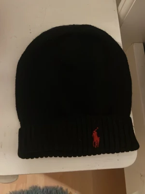 Svart mössa från Polo Ralph Lauren - Snygg svart mössa från Polo Ralph Lauren med röd broderad logga framtill. Klassisk ribbstickad kant och mjukt material som sitter skönt på huvudet. Perfekt accessoar för kalla dagar och enkel att matcha med olika outfits.