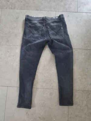 Mörkgrå skinny jeans - Säljer ett par mörkgrå skinny jeans med klassisk femficksdesign och smala ben. Jeansen har en diskret detalj på bakfickan och tryckt logga i midjan. Perfekta för dig som gillar en enkel men snygg stil. Bredden på dondup jeansen är 40 cm och från skrevet till nedåt är det 65cm alltså innre längden. Men ifall man mäter topp till botten på jeansen så är det 90cm