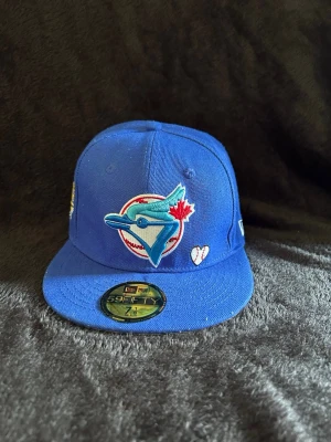 Blå Toronto Blue Jays keps - Säljer en blå fitted-keps med Toronto Blue Jays-logga framtill och broderad World Series 1992-patch på sidan. Kepsen har platt skärm och klassisk 59FIFTY-stil. Materialet är bomull och kepsen har detaljer i rött, vitt och blått.