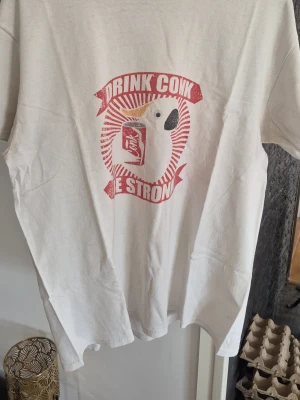 Vit t-shirt med papegojtryck - Vit t-shirt med ett tryck av en papegoja som håller en läskburk och texten 'Drink Coke Be Strong' i rött. T-shirten har korta ärmar och en klassisk passform. Perfekt för dig som gillar roliga och unika motiv.