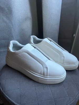 Vita slip-on sneakers utan snörning - Stilrena vita slip-on sneakers med elastisk panel över vristen för enkel påtagning. Skorna har en minimalistisk design utan snörning och är tillverkade i syntetmaterial med gummisula. Perfekta för en clean och avslappnad look. Storlek 39 men stora på mig som brukar ha 39.