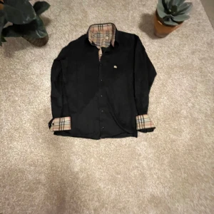 Svart skjorta från Burberry - Snygg svart skjorta från Burberry med klassiskt rutigt mönster på insidan av kragen och ärmsluten. Skjortan har långa ärmar och knappar framtill. Perfekt för en stilren look.