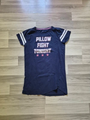 Mörkblå nattlinne med tryck - Mörkblå nattlinne i t-shirtmodell med vita ränder på ärmarna och texten 'Pillow Fight Tonight' framtill. Kortärmad och rund halsringning, tillverkad i mjukt bomullsliknande material. Perfekt för mysiga kvällar och sömn.