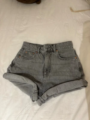 Gråa högmidjade jeansshorts - Säljer ett par gråa högmidjade jeansshorts med uppvikta benslut och klassisk femficksdesign. Shortsen har bälteshällor och stängs med knapp och dragkedja. Perfekta för en avslappnad och trendig look.