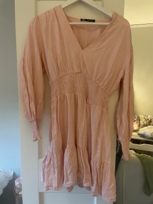 Ljusrosa omlottklänning med volang - Söt ljusrosa klänning med omlott upptill, smock i midjan och långa ärmar. Klänningen har volangdetaljer nedtill och är tillverkad i ett mjukt, tunt tyg som passar perfekt till varmare dagar.
