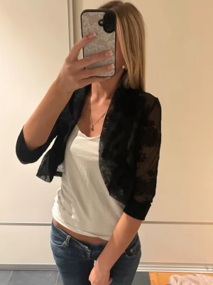 Svart kort spetscardigan - Snygg svart cardigan i kort modell med trekvartsärmar. Tillverkad i genomskinlig spets med blommigt mönster som ger en elegant touch. Perfekt att slänga över ett linne eller topp för en trendig look.