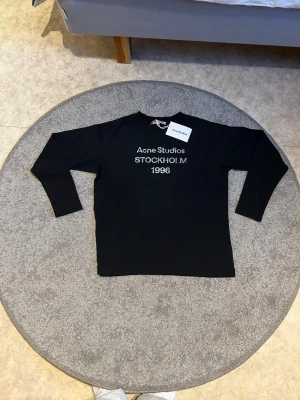 Svart långärmad t-shirt Acne Studios - Svart långärmad t-shirt från Acne Studios med tryck i vitt framtill där det står 'Acne Studios STOCKHOLM 1996'. Klassisk rak passform och rund hals. Perfekt för dig som gillar stilrena och trendiga plagg.