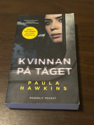 Kvinnan på tåget pocketbok - Spännande pocketbok 'Kvinnan på tåget' av Paula Hawkins. Omslaget är mörkt med en bild på en kvinna i ett tågfönster och text i vitt och gult. Boken är utgiven av Massolit Pocket och är en bästsäljare som även blivit film.