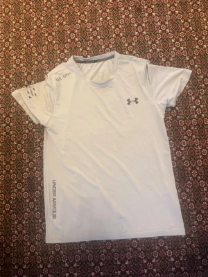 Vit tränings-tshirt från Under Armour - Snygg vit tränings-tshirt från Under Armour med reflekterande logga och detaljer. T-shirten har rund hals, korta ärmar och är tillverkad i ett lätt och snabbtorkande material som passar perfekt till träning och löpning. Storlek M passar S