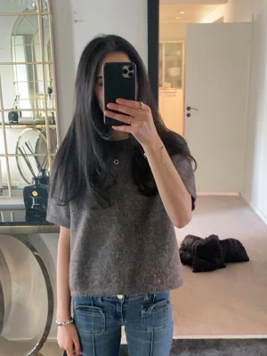 Mörkgrå kortärmad ulltopp - Säljer en enkel och stilren mörkgrå topp i ull med korta ärmar och rak passform. Perfekt att matcha med jeans eller kjol för en avslappnad look. Toppen har en mjuk och lite luddig yta som ger en mysig känsla.