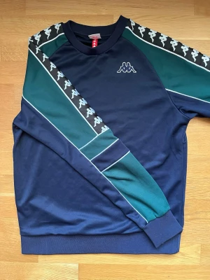 Blågrön stickad tröja från Kappa - Snygg stickad tröja från Kappa i marinblått och mörkgrönt med vita detaljer. Tröjan har klassiska Kappa-loggor längs ärmarna och ribbade muddar vid ärmslut och nederkant. Perfekt för en sportig och avslappnad stil.
