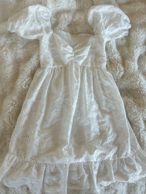 Vit kort klänning med puffärm - Söt vit kort klänning med puffärmar och broderade blommor. Klänningen har volangkant nertill, smock upptill och v-ringad urringning. Perfekt för sommarens alla tillfällen och ger en romantisk vibe.