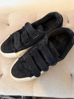 Svarta sneakers med kardborreband - Säljer ett par svarta sneakers i manchestertyg med tre breda kardborreband och vit sula. Skorna har en klassisk låg modell och en cool retrostil som passar till det mesta. Perfekta för dig som gillar enkel design och bekväma skor.