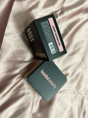 Huda Beauty Easy Bake Cherry Blossom - Säljer ett Huda Beauty Easy Bake Loose Baking & Setting Powder i färgen Cherry Blossom Cake. Kommer i en fyrkantig burk med svart lock och rosa text. Pudret har en ljusrosa ton och är perfekt för att fixera makeup och ge en mjuk finish.