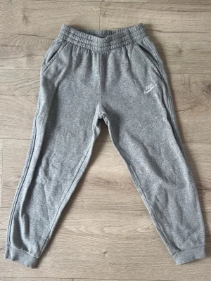 Gråa mjukisbyxor från Nike - Snygga gråa mjukisbyxor från Nike med resår i midjan och muddar vid bensluten. Byxorna har två sidofickor och en liten vit Nike-logga på vänster ben. Tillverkade i mjukt och bekvämt material, perfekta för chill dagar.