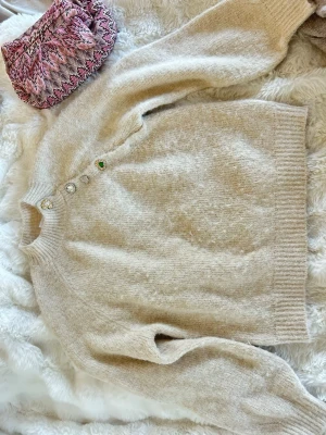 Beige stickad tröja med dekorativa knappar - Mysig beige stickad tröja med ribbad hals, ärmslut och nederkant. Tröjan har unika dekorativa knappar i olika former och färger längs axeln, bland annat en grön och flera glittriga stenar. Perfekt för dig som vill ha något extra i garderoben.