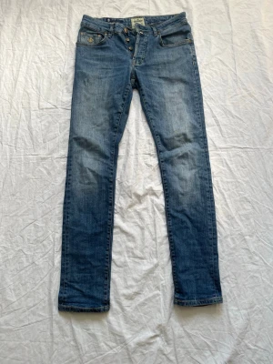 Blå jeans från Morris, rak modell - Snygga blå jeans från Morris i rak modell med knäppning fram och klassiska fem fickor. Jeansen har guldfärgade detaljer och en liten broderad logga på myntfickan. Tillverkade i slitstark denim som passar perfekt till din vardagsstil.