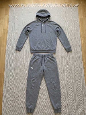  Under Armour set - Mysigt grått loungewear-set från Under Armour med hoodie och matchande byxor. Hoodien har dragsko och magficka, medan byxorna har resår i midjan och muddar vid bensluten. Perfekt för chill dagar hemma eller när du vill vara bekväm.