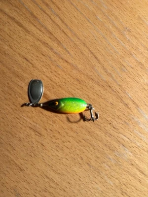 Grön och gul fiskedrag med sked - Säljer ett fiskedrag i grönt och gult med svart rygg och orange mage. Draget är en lotto spinnare. Draget har en metallsked framtill och trekrok baktill. Perfekt för dig som gillar fiske och vill ha ett färgglatt bete i lådan.