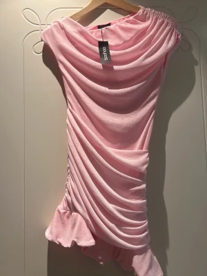 Babyrosa draperad klänning Boohoo - Supersnygg babyrosa kortklänning från Boohoo med draperad design och asymmetrisk volangkant. Klänningen har en axel och är tillverkad i ett mjukt, stretchigt material som ger en smickrande passform. 