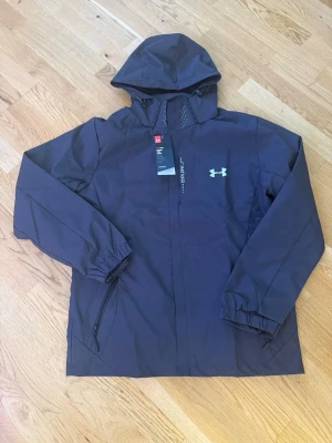 Svart vindjacka från Under Armour - Svart sportjacka från Under Armour med huva. Den passar M/L