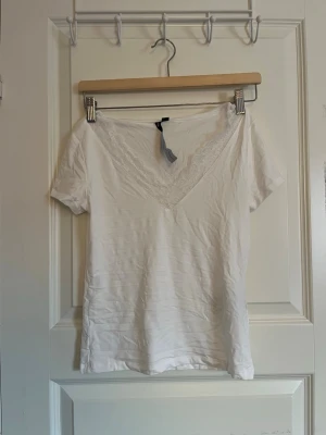 Vit t-shirt med spetsdetalj - Säljer en vit t-shirt med v-ringning och snygg spetsdetalj längs halsen. T-shirten har korta ärmar och är gjord i mjukt material. Har inte kommit till användning 