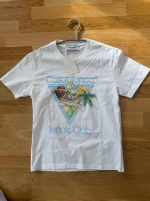 Vit t-shirt från Casablanca - Säljer en vit t-shirt från Casablanca med färgglad grafik och texten 'Casablanca Tennis Club' på bröstet. T-shirten har rund hals, korta ärmar och är gjord i mjuk bomull. Perfekt för dig som gillar stilrena plagg med coola tryck.