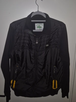 Svart vindjacka och byxa från Lacoste - Snygg svart Tracksuit från Lacoste med dragkedja framtill och två fickor med gula detaljer. Jackan har hög krage och diskret Lacoste-logga på bröstet. Perfekt för blåsiga dagar och sportiga outfits.