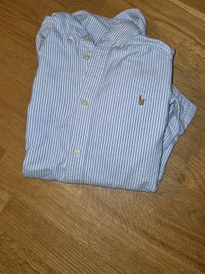 Blårandig skjorta från Ralph Lauren - Klassisk blårandig skjorta från Ralph Lauren med button-down krage och broderad logga på bröstet. Skjortan är tillverkad i mjuk bomull och har vita knappar framtill. Perfekt för en stilren och avslappnad look.