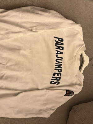 Vit sweatshirt från Parajumpers - Vit sweatshirt från Parajumpers med stort svart tryck på bröstet och orange tryck på insidan av nederkanten. Långärmad med ribbade muddar och patch på ena ärmen. Snygga orange sömmar vid axlarna. Perfekt för chill dagar.