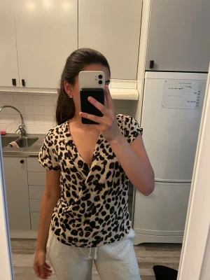 Leopardmönstrad omlottopp från Zara - Snygg omlottopp från Zara med leopardmönster i svart och beige. Toppen har djup v-ringning och korta ärmar, perfekt för att sticka ut lite extra. Tillverkad i mjukt och lätt material som känns skönt mot huden.