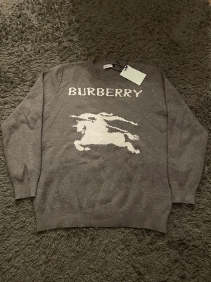 Grå stickad tröja från Burberry - Snygg grå stickad tröja från Burberry med vit logga och ikonisk ryttare framtill. Tröjan har rund halsringning och ribbade muddar vid ärmslut och nederkant. Perfekt för dig som gillar klassisk design med en modern twist.