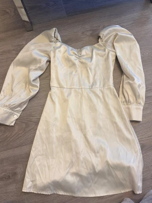 Ljus kort klänning med puffärm - Snygg ljus kort klänning med puffiga långärmar och markerad midja. Klänningen har en lätt glansig yta och smock i ryggen för skön passform. Perfekt för dig som gillar romantisk och trendig stil.